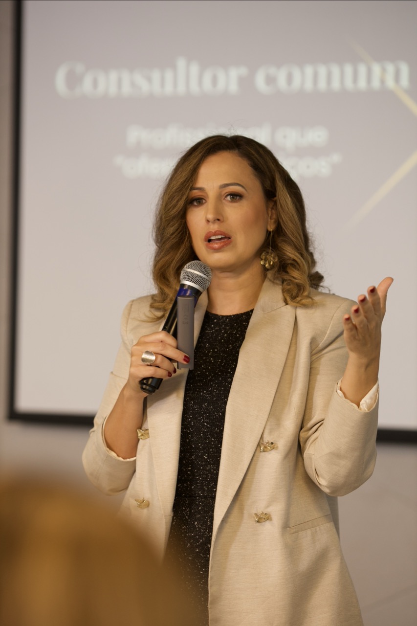 Juliana Paes Garcia no palco de um workshop, microfone na mão, blazer bege, expressão de autoridade